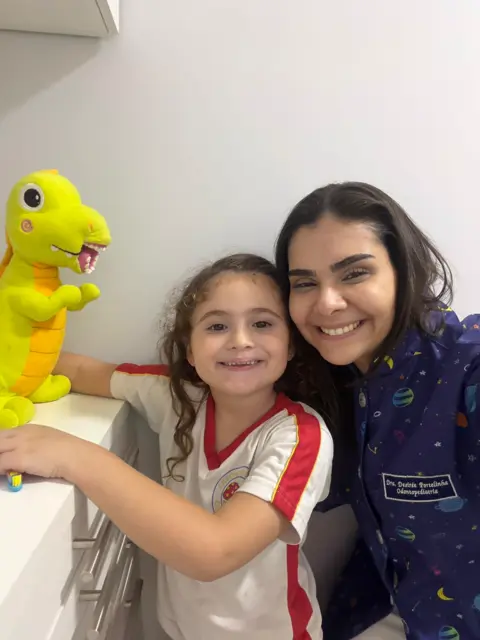 A Dra. Desirée e uma menina sorrindo ao lado do Dino, a pelúcia de dinossauro amarela com dentes.
